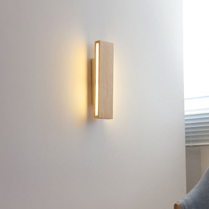 Brandende verticale houten wandlamp aan een lichte muur, met warm licht en een raam met jaloezie en stoel ernaast. 15