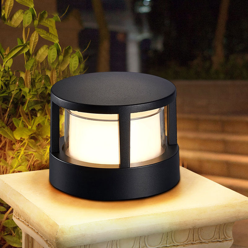 Ronde zwarte buitenlamp met vlak dak en gloeiend melkglas, geplaatst op een stenen sokkel in een verlichte tuin. 1