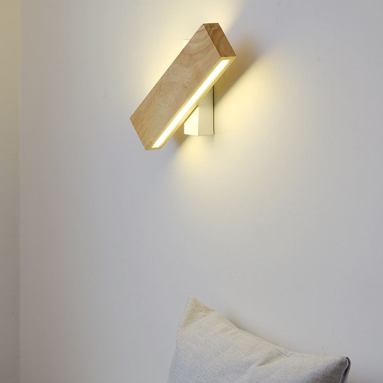 Houten ledwandlamp met witte bevestiging, schuin gedraaid en brandend, aan een lichte muur boven een kussen. 1