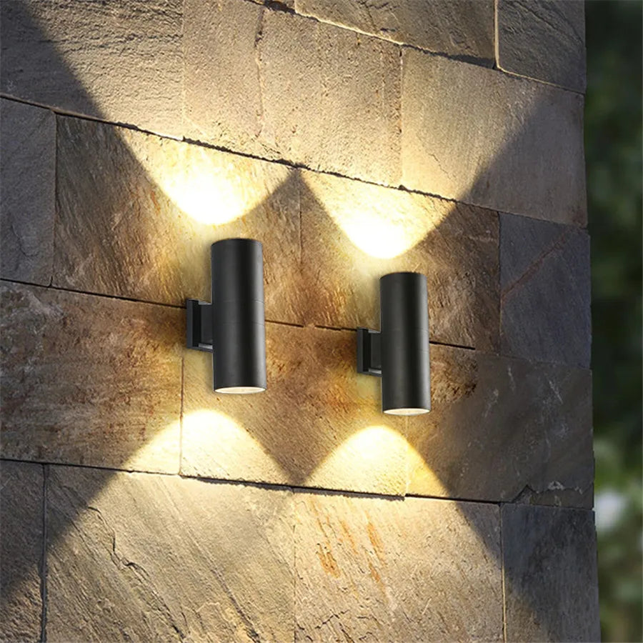 Twee zwarte cilinderlampen op een stenen buitenmuur die warm licht omhoog en omlaag werpen en zo lichtwaaiers op de natuursteen tekenen. 2