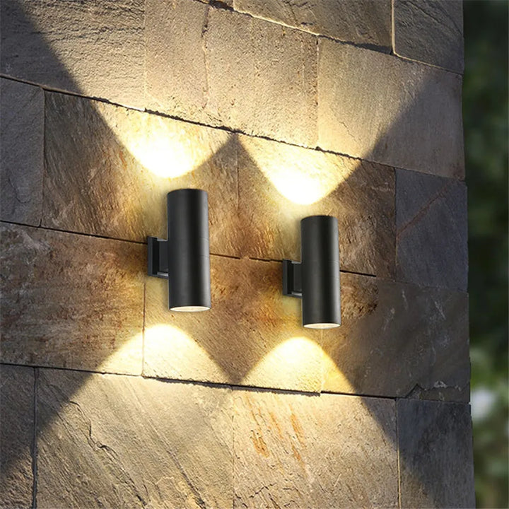 Twee zwarte cilinderlampen op een stenen buitenmuur die warm licht omhoog en omlaag werpen en zo lichtwaaiers op de natuursteen tekenen. 2