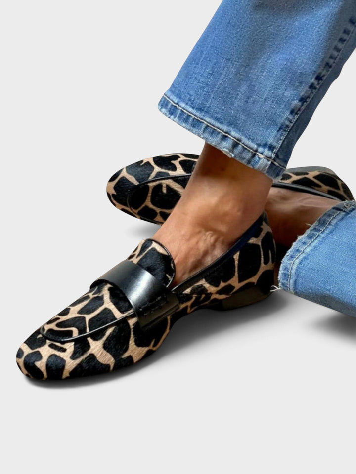 Gedragen loafers met dierenprint en zwarte leren band, zijaanzicht met gekruiste voeten, gecombineerd met een blauwe jeans op lichte ondergrond. 1