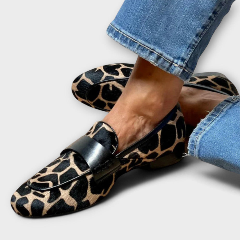 Close-up van de gedragen loafers met zwarte leren band en gevlekte beige-zwarte print, de voeten gekruist naast een jeansbroekspijp. 2