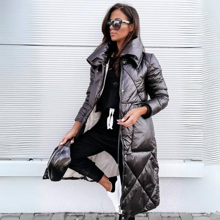 Vrouw met zonnebril draagt de zwarte, glanzende gewatteerde lange jas open, met zwarte outfit, witte sneakers en een zwarte heuptas. 1