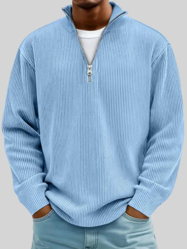 Model draagt de geribbelde trui in lichtblauw met halve rits, een wit shirt eronder en handen in de zakken van een spijkerbroek. 1