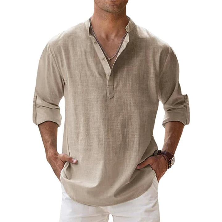 Man draagt het losvallende Henley overhemd in beige met opgerolde mouwen, handen in de zakken van een witte broek. 1