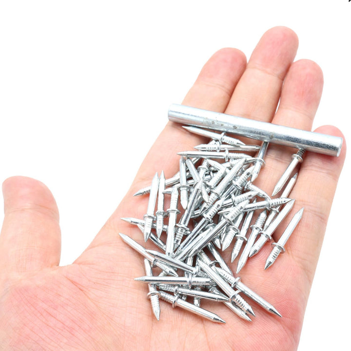 Een hand houdt een hoop zilverkleurige dubbele spijkers vast met daarbovenop een metalen hulsje om de nagels in te zetten. 1