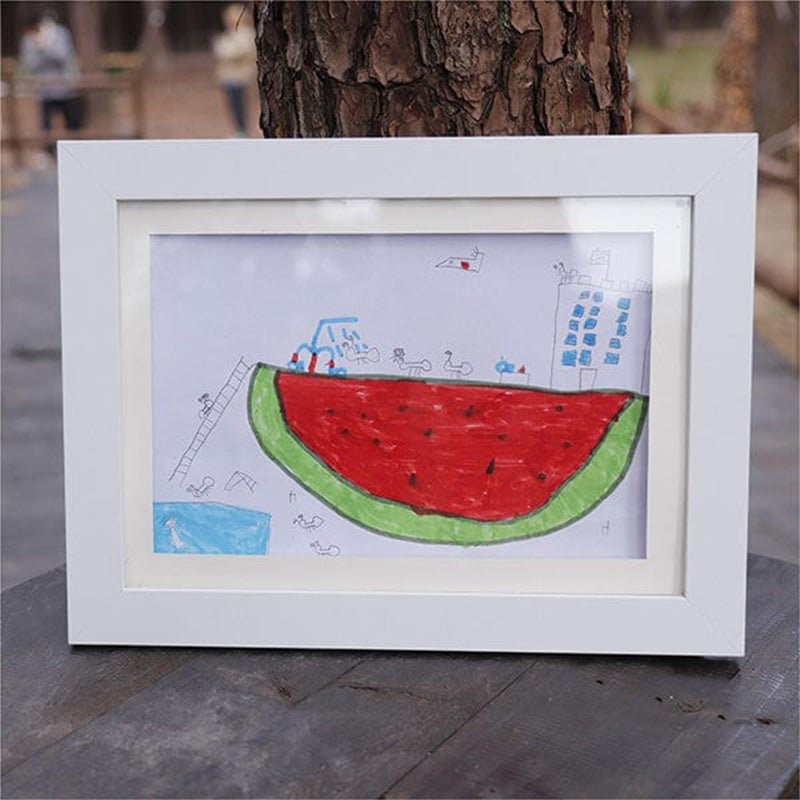 Witte lijst staat buiten op een houten tafel en toont een kindertekening van een grote rode watermeloenpartj met een gebouw en trap. 21