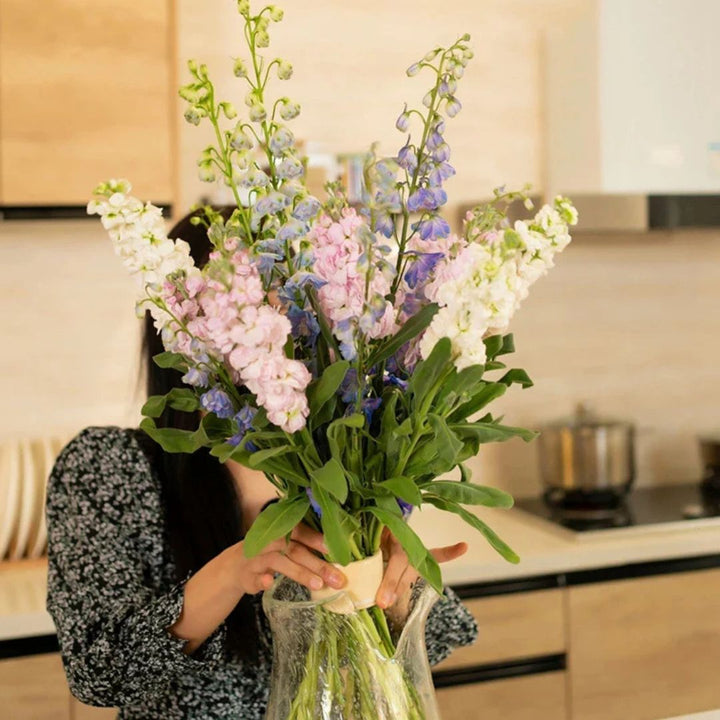 Vrouw in een keuken houdt een hoog, fris boeket roze, witte en blauwe bloemen vast, met de inzet in de glazen vaas. 2