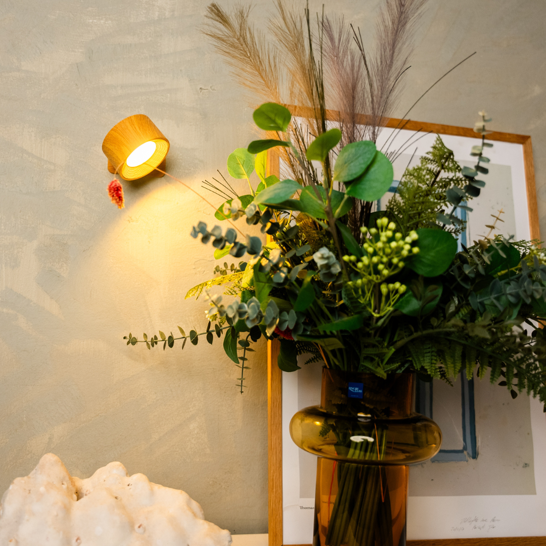 Houtkleurige wandlamp brandt naast een vaas met groene bladeren en pampasgras tegen een grijsgroene muur. 6