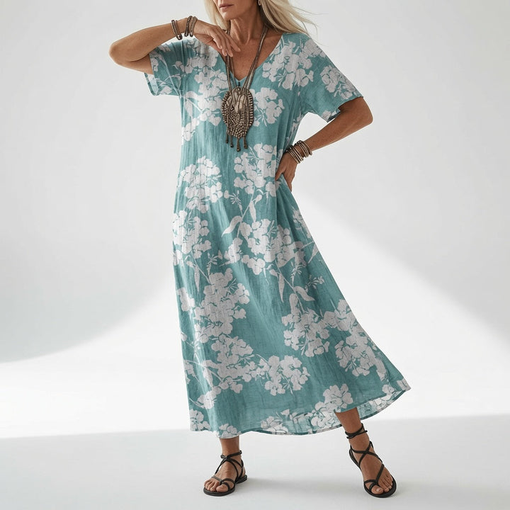 Model draagt de turquoise jurk met een witte bloemenprint, een opvallende hanger en zwarte sandalen met gekruiste bandjes. 4