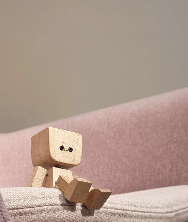 Klein houten figuurtje met glimlachend gezichtje zit op de rand van een roze bank. 4