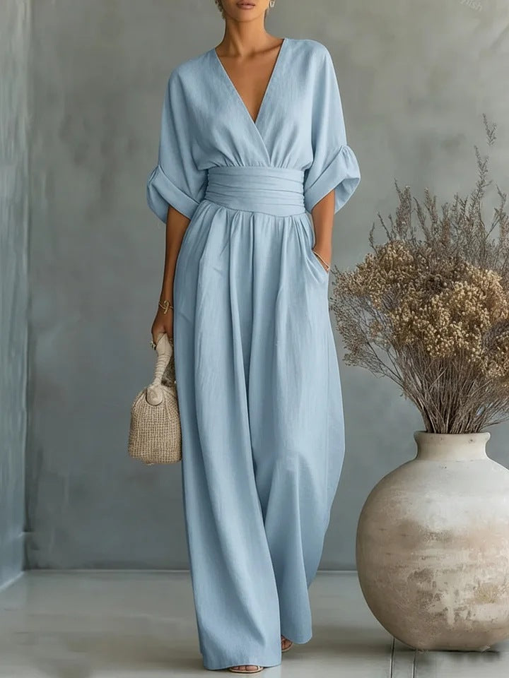 Dezelfde jumpsuit in lichtblauw, frontaal gedragen door een model met rieten tasje naast een vaas met droogbloemen. 2
