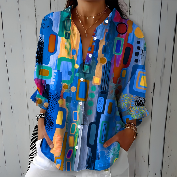 Model draagt de losse blouse in blauwtinten met kleurrijk abstract patroon, handen in de zakken van een witte broek, vooraanzicht. 1