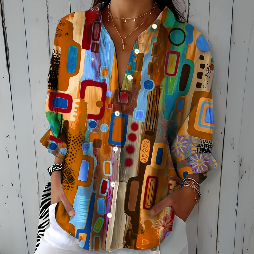Model draagt de blouse in oranje en bruine tinten met blauwe accenten en een abstract patroon, handen in de broekzakken. 3