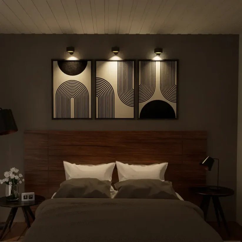 Drie zwarte wandlampen boven drie abstracte lijnkunstwerken in een slaapkamer boven een houten hoofdbord, 's avonds aan. 5