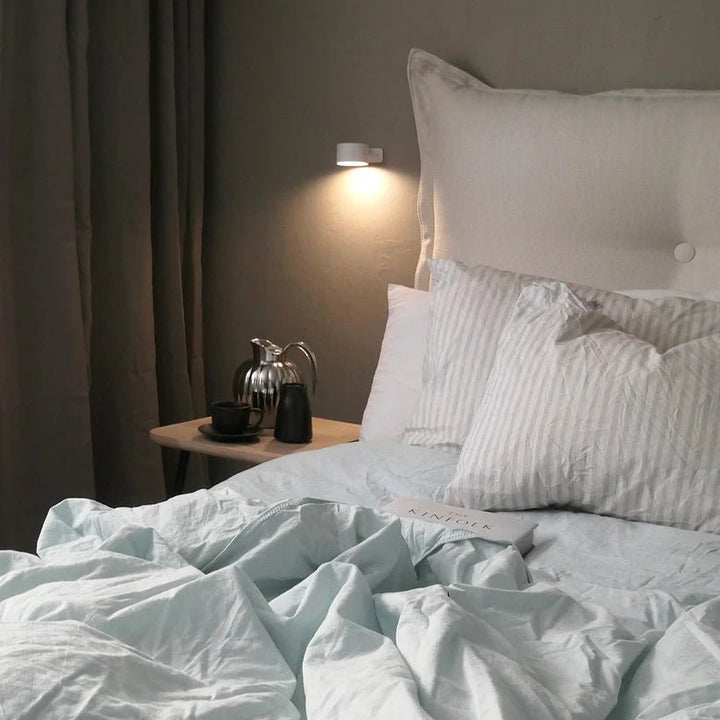 Kleine witte wandlamp naast het bed geeft zacht licht boven een nachtkastje met kan en kopje. 7