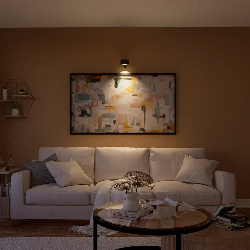 Zwarte wandlamp boven een ingelijst abstract schilderij verlicht het kunstwerk in een woonkamer met crème bank. 8