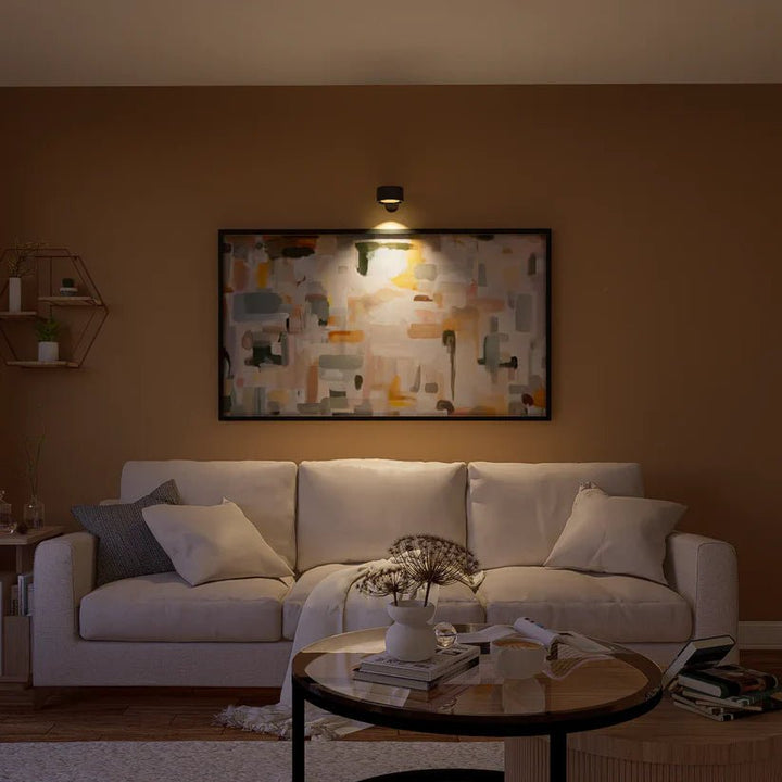 Zwarte wandlamp boven een ingelijst abstract schilderij verlicht het kunstwerk in een woonkamer met crème bank. 8