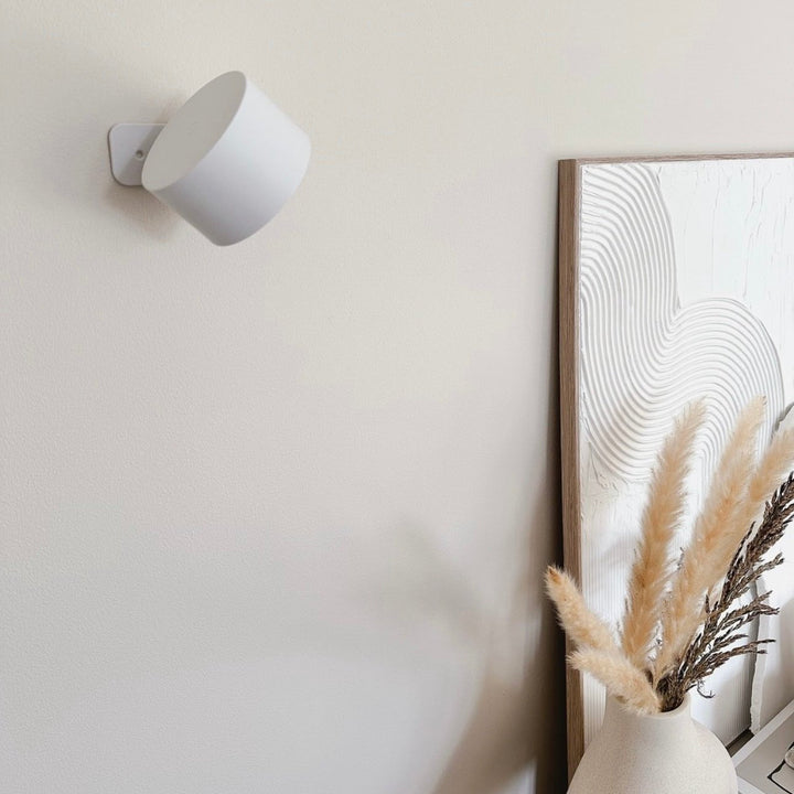 Witte wandlamp, uitgeschakeld en schuin gedraaid, aan een beige muur naast een kunstwerk en pampasgras in een vaas. 17