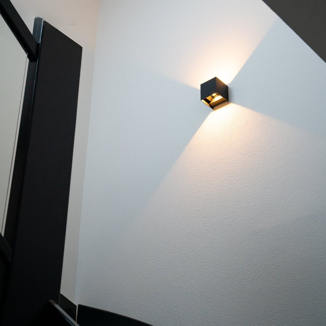 Zwarte kubuslamp aan een witte, gestructureerde trapmuur, gezien van onderaf, met warm licht dat omhoog en omlaag uitwaaiert. 2