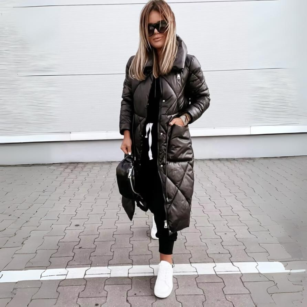 Vrouw met blond haar draagt de zwarte lange gewatteerde jas dichtgeritst, met een hand in de zak, zwarte tas en witte sneakers. 5
