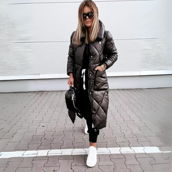 Vrouw met blond haar draagt de zwarte lange gewatteerde jas dichtgeritst, met een hand in de zak, zwarte tas en witte sneakers. 5