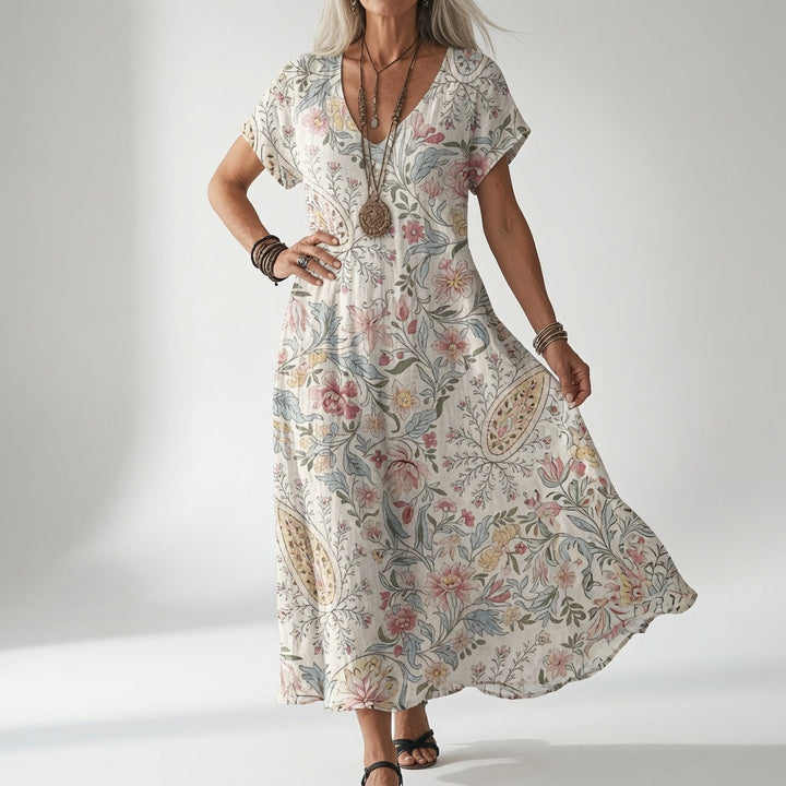 Vrouw draagt de jurk in crèmewit met een fijn roze, blauw en geel bloemen- en paisleymotief, gecombineerd met houten kettingen en sandalen. 1