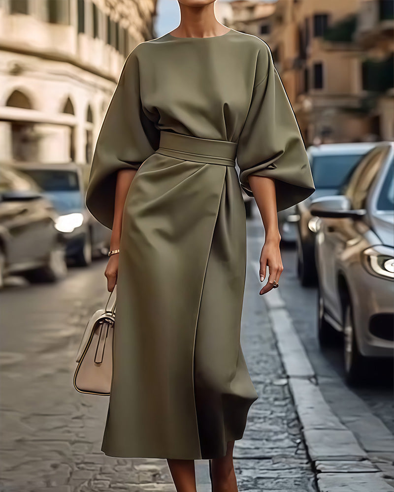 Detail van de jurk in groenachtig olijfgrijs met ruime halflange mouwen en brede ceintuur, gedragen met een beige handtas. 2
