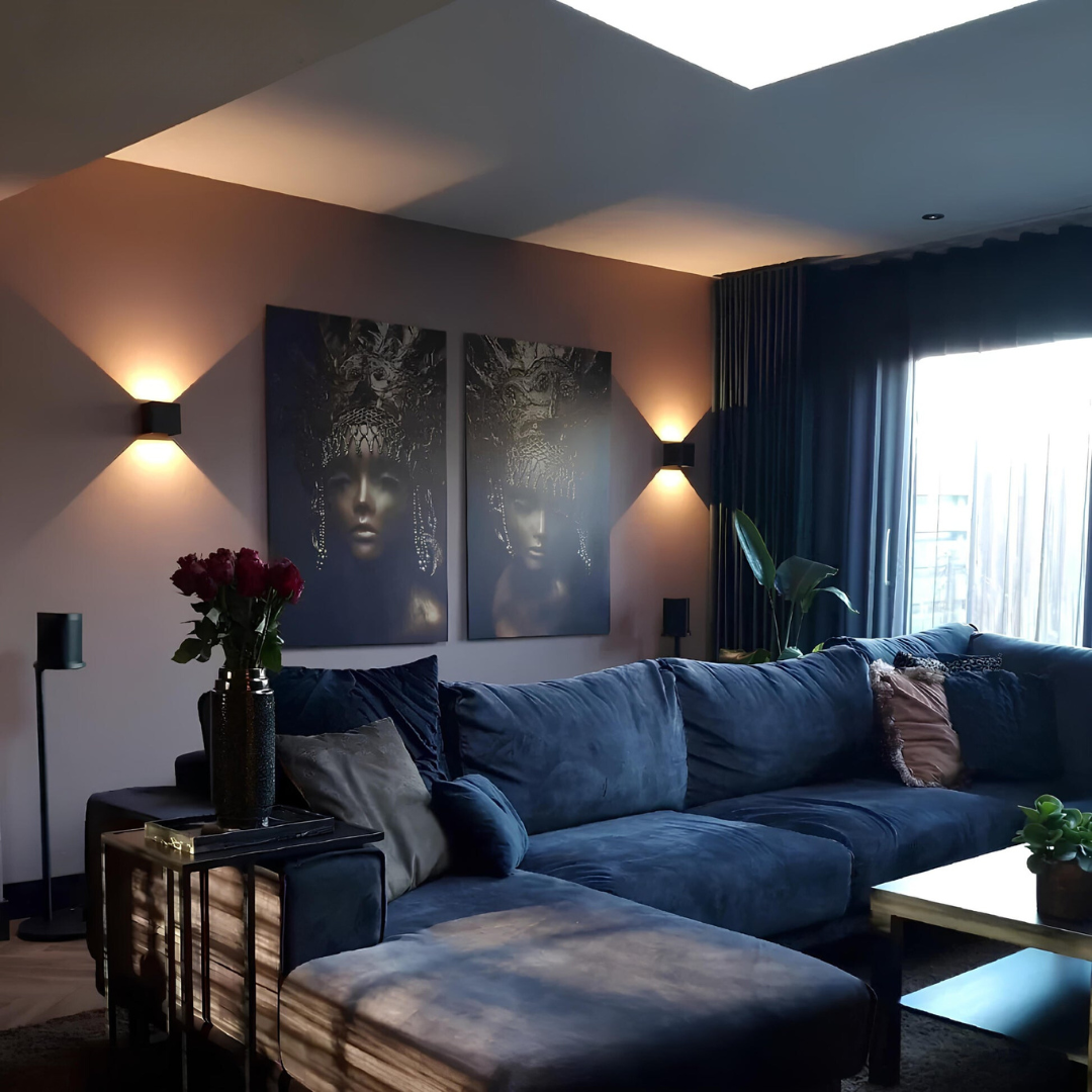 Zwarte wandlampen aan weerszijden van twee kunstwerken boven een blauwe hoekbank in een woonkamer. 9