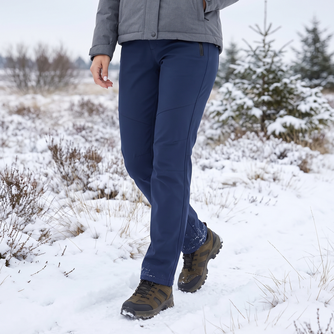 Vrouw in marineblauwe broek en wandelschoenen loopt door een besneeuwd heidelandschap met sparren 1