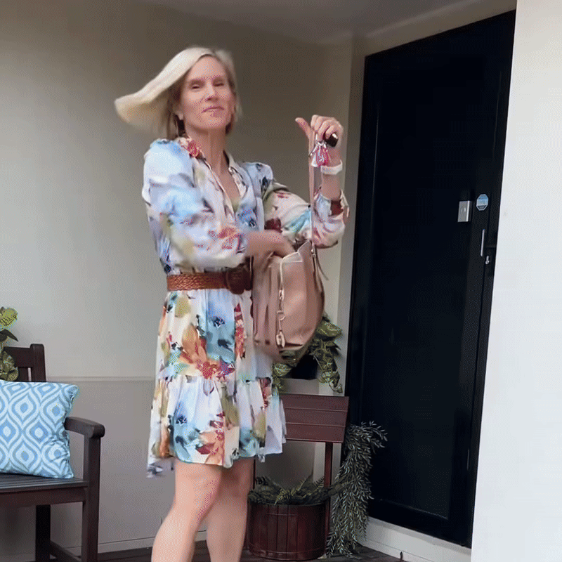 Blonde vrouw in een bloemenjurk met riem en handtas schudt haar gestylde haar bij een zwarte voordeur. 8