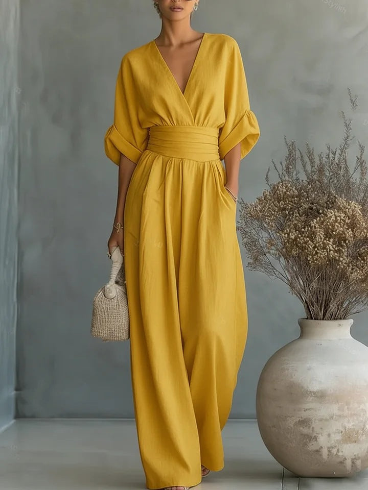 Dezelfde jumpsuit in mosterdgeel, frontaal gedragen met hand in de zak, naast een vaas met droogbloemen. 3