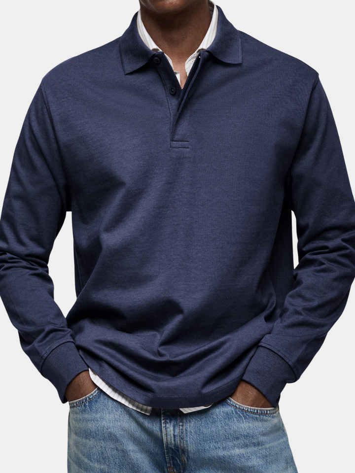 Man draagt het marineblauwe poloshirt met knoopsluiting en ribboorden, handen in de zakken van een lichte spijkerbroek, close-up van de voorkant. 1