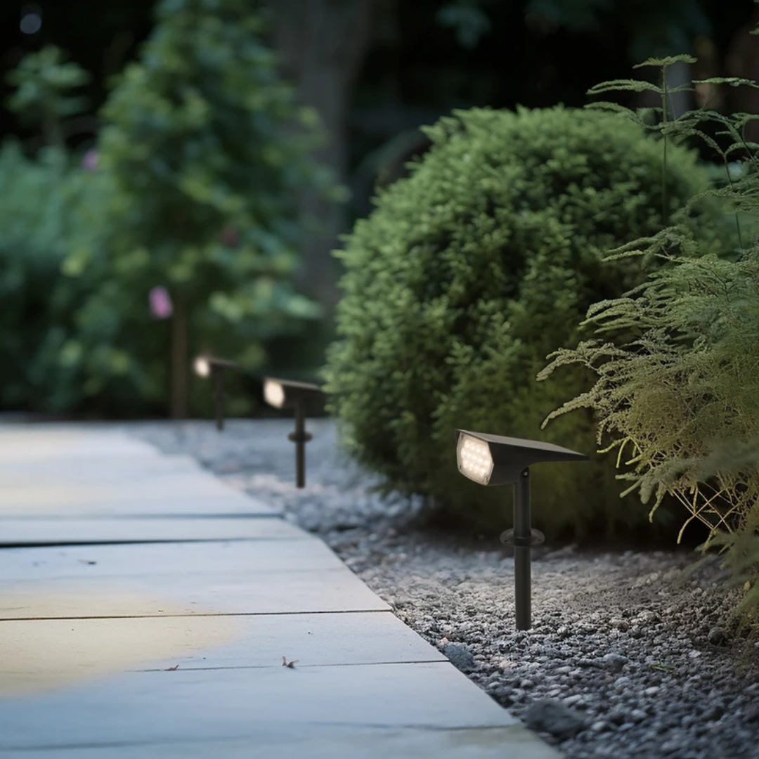 Drie zwarte zonnespots met grondpen langs een tuinpad van grind en tegels, 's avonds naast een ronde struik in warm licht. 1