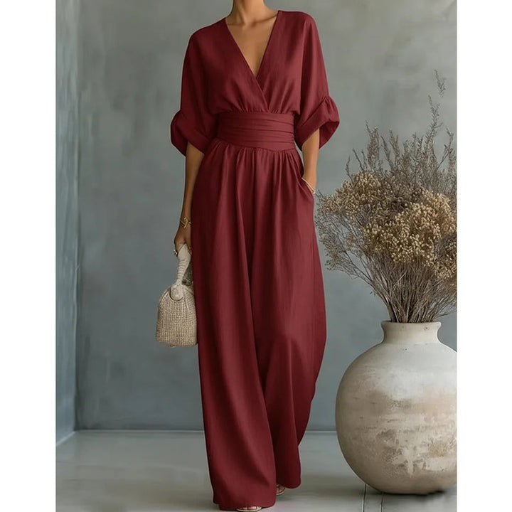 Wijnrode jumpsuit met V-hals, brede taillestrook en wijde pijpen, frontaal gedragen naast een vaas met droogbloemen. 8