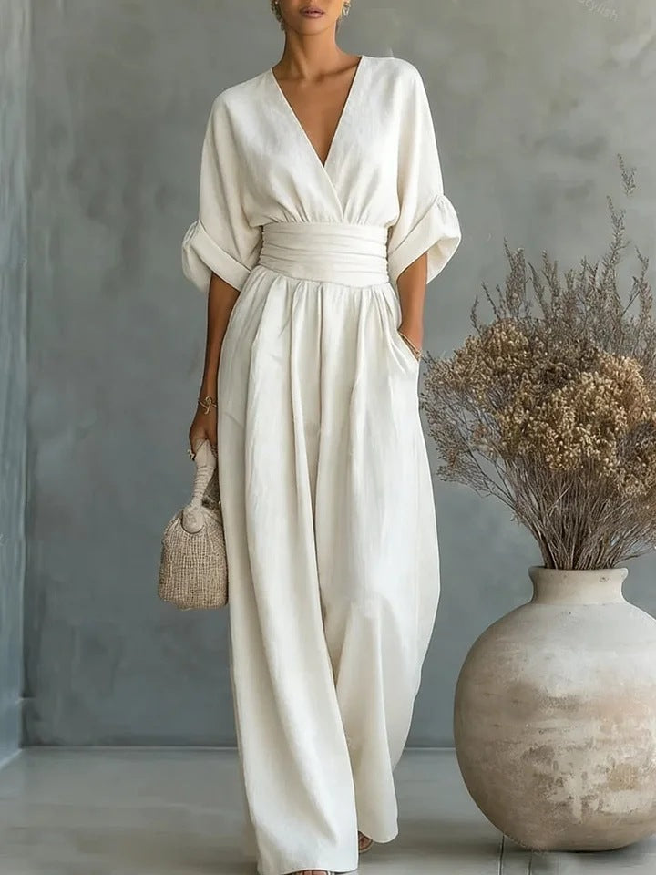 Model in crèmewitte jumpsuit met wikkelhals en brede taillestrook, naast een grote vaas met droogbloemen, rieten tasje in de hand. 1