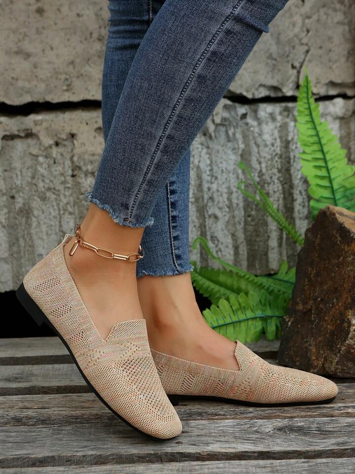 Zijaanzicht van beige gebreide instapschoenen met zwarte zool, gedragen met spijkerbroek op een houten vloer naast groene varens. 4