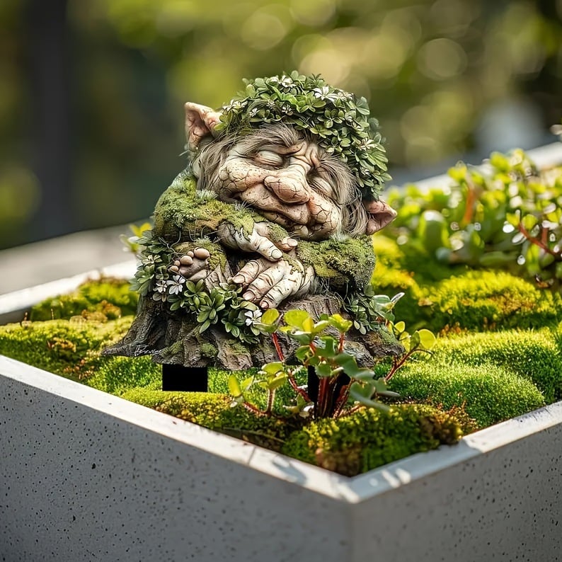 Slapend boomwezen met bladerkrans en gevouwen handen op een boomstronk, geplaatst in een betonnen plantenbak vol mos en vetplanten. 4