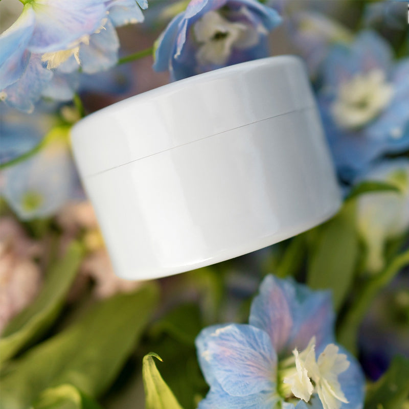 Witte ronde inzet met deksel, schuin tussen blauwe en roze bloemen en groene bladeren gelegd. 9