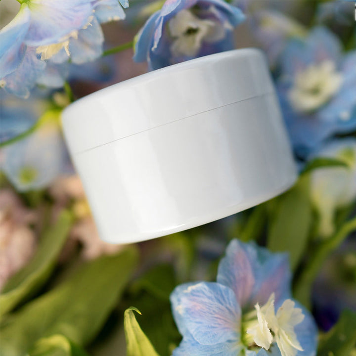 Witte ronde inzet met deksel, schuin tussen blauwe en roze bloemen en groene bladeren gelegd. 15