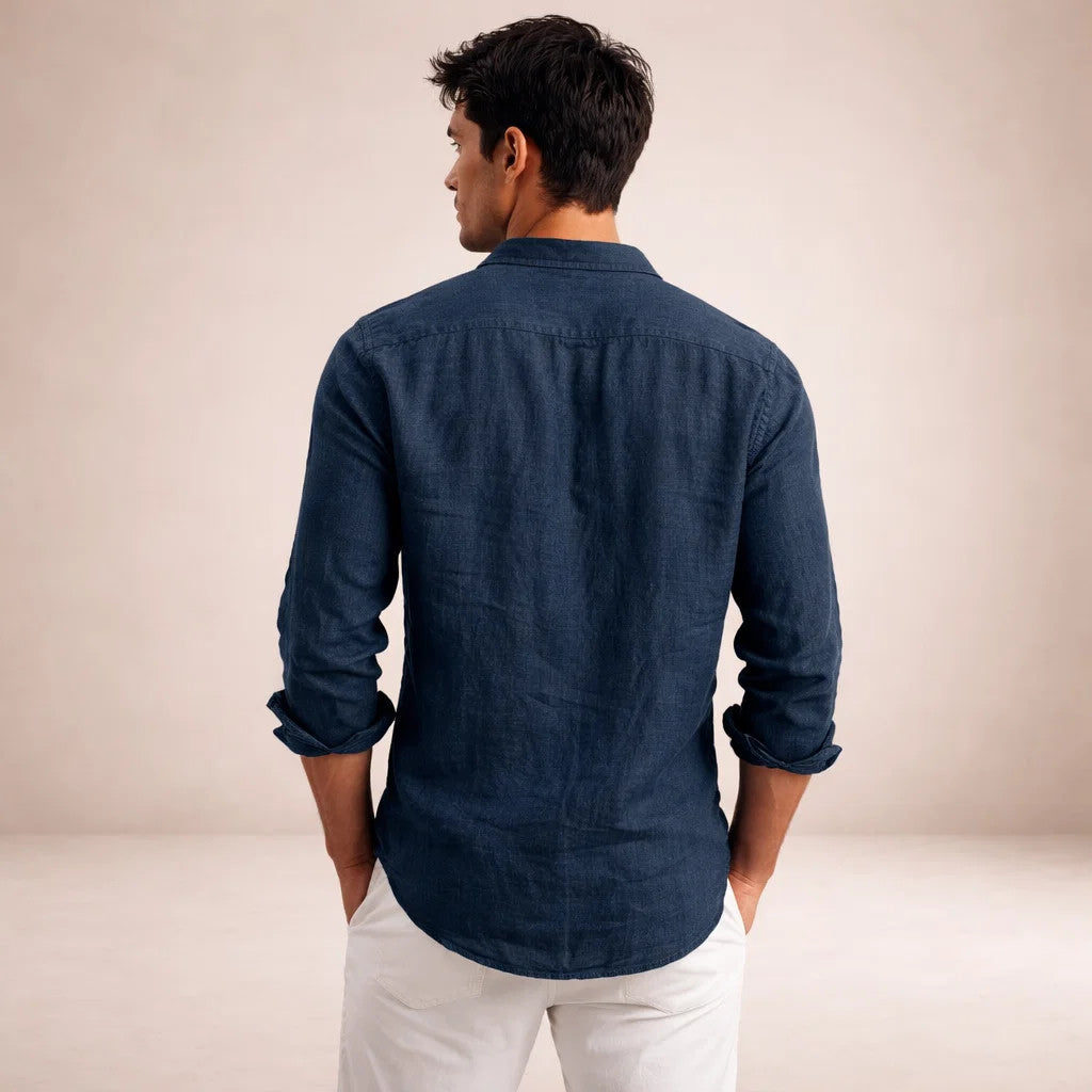 Achterkant van het marineblauwe linnen overhemd aan een model dat zijn hoofd draait, met handen in de zakken van een witte broek. 13