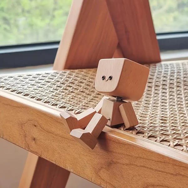 Houten figuurtje met lachend gezichtje zit op de rand van een houten stoel met rotanzitting, voor een raam. 11