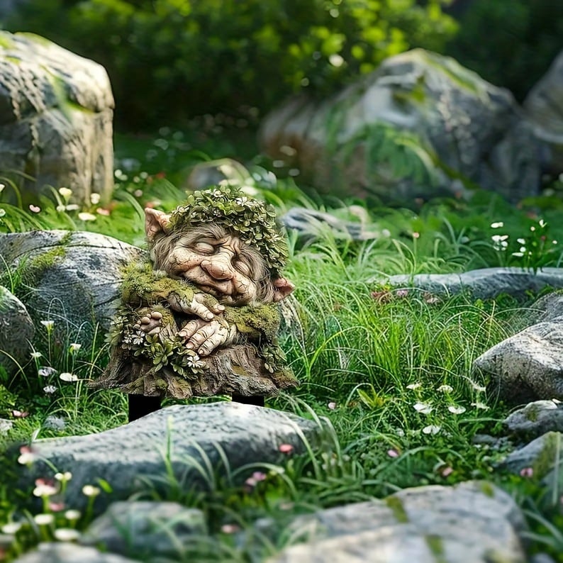 Slapend boomwezen met bladerkrans en gevouwen handen, tussen rotsen, hoog gras en kleine bloemetjes in een natuurlijke omgeving. 16