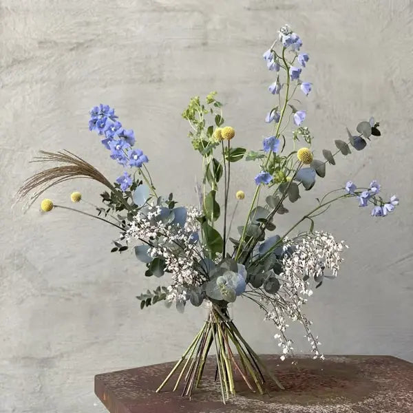 Los gebonden boeket met blauwe bloemen, gele ballen en eucalyptus, rechtopstaand op een roestige ondergrond tegen een grijze muur. 17