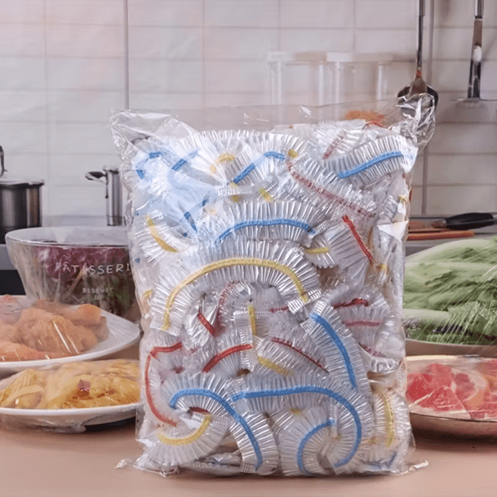 Grote doorzichtige zak vol elastische afdekkappen met blauwe, gele en rode randen, staand op een keukenblad tussen afgedekte schalen. 1