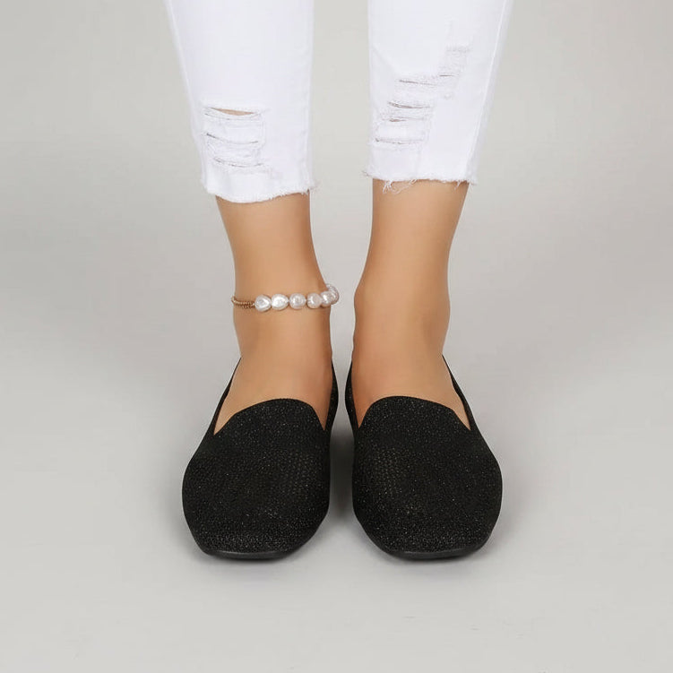 Voorkant van zwarte instapschoenen met vierkante neus, gedragen met witte gescheurde jeans en een parelenkelbandje op een lichtgrijze achtergrond. 2
