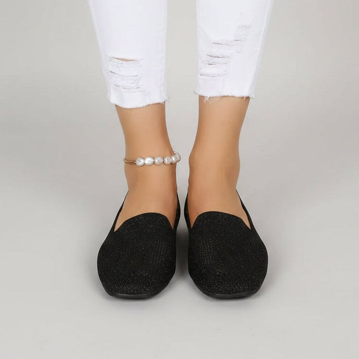 Voorkant van zwarte instapschoenen met vierkante neus, gedragen met witte gescheurde jeans en een parelenkelbandje op een lichtgrijze achtergrond. 2