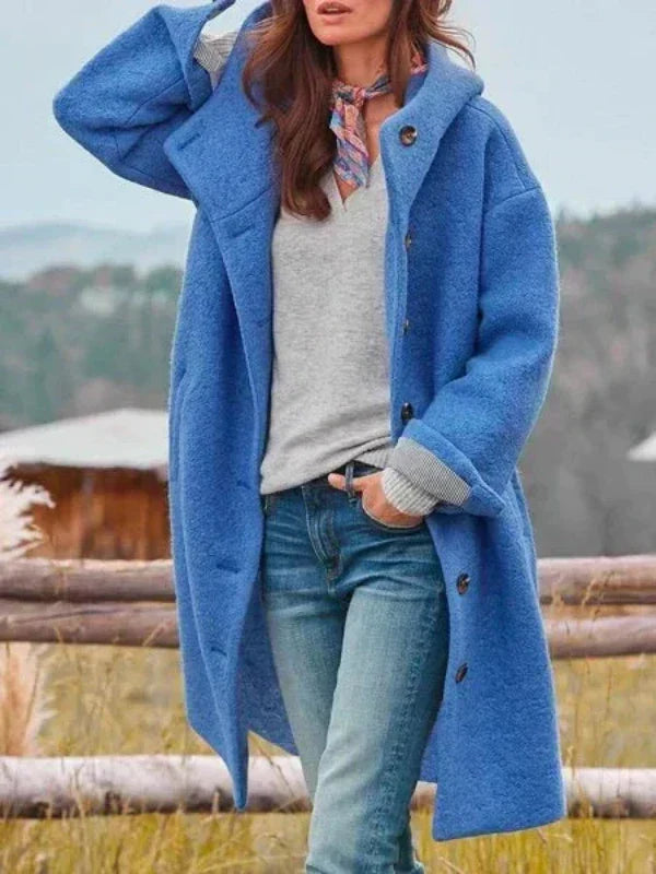 Vrouw draagt de lange blauwe jas met capuchon open over een grijze trui, jeans en een kleurige sjaal. 4