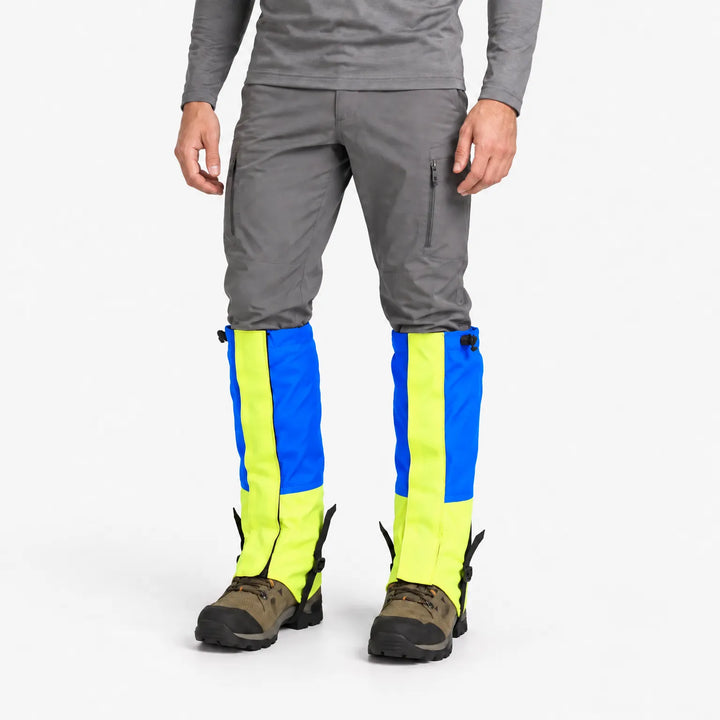 Man in grijze broek draagt blauwe gamaschen met neongele panelen en zwarte bandjes onder de wandelschoenen, tegen witte achtergrond. 2